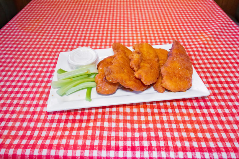 A white plate of golden-brown chicken tenders, celery sticks, and ranch dressing on a red and white checkered tablecloth.