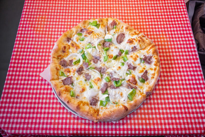 Whole pizza with sausage, green peppers, and onions on a red and white checkered tablecloth.