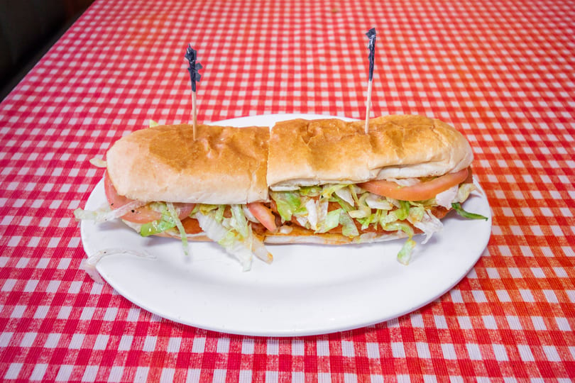 A long submarine sandwich with lettuce and tomato on a white plate, on a red and white checkered tablecloth.