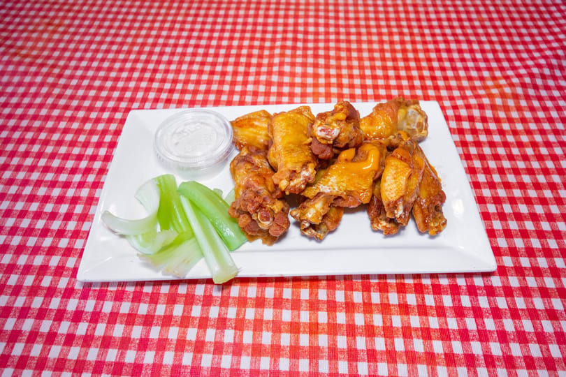 A plate of buffalo wings with celery sticks and ranch dressing on a red and white checkered tablecloth.